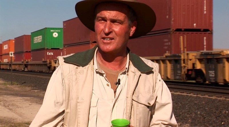 Gordon in a wide-brimmed hat beside a passing freight train.